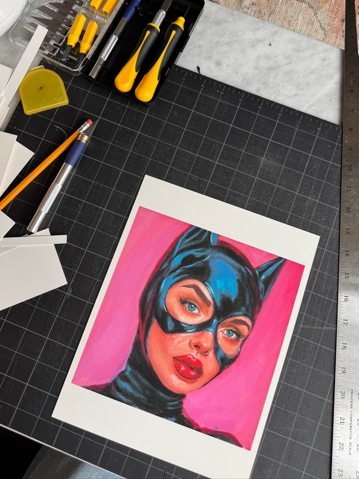 A colorful art print of a woman in a black cat costume against a bright pink background, displayed on a black cutting mat surrounded by craft tools and materials.