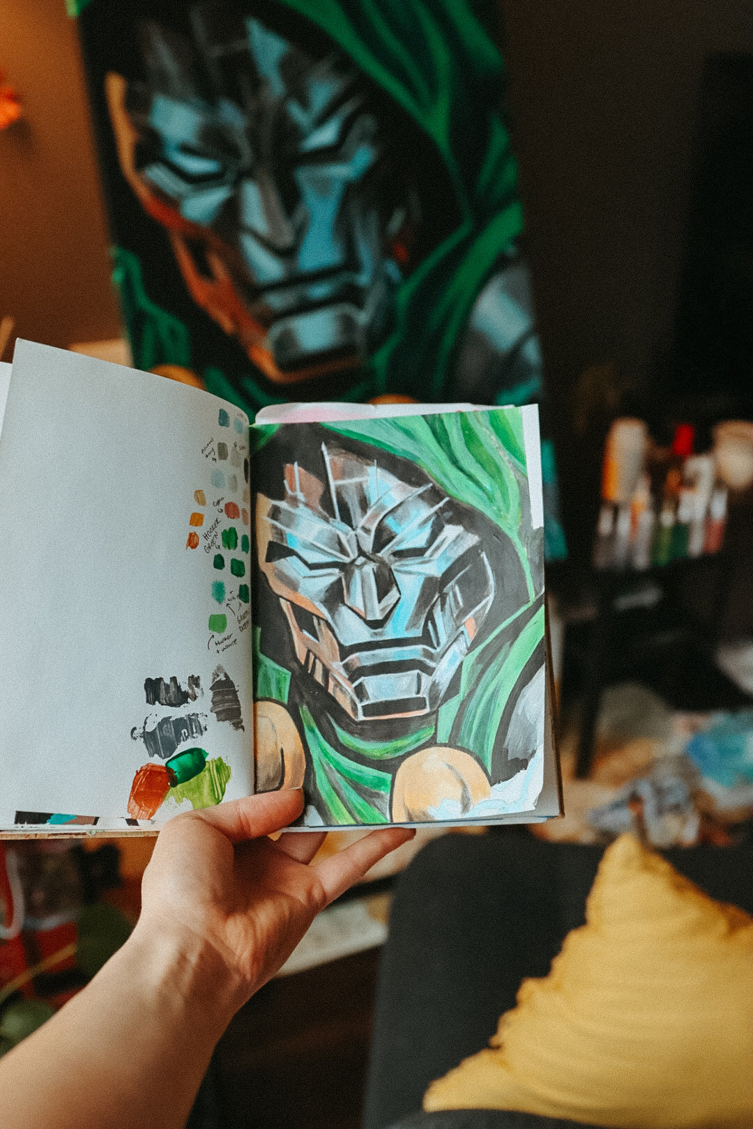 A hand holds an open sketchbook showing a colored illustration of a metallic blue and green character mask, with a larger matching artwork hanging on the wall in the blurred background.