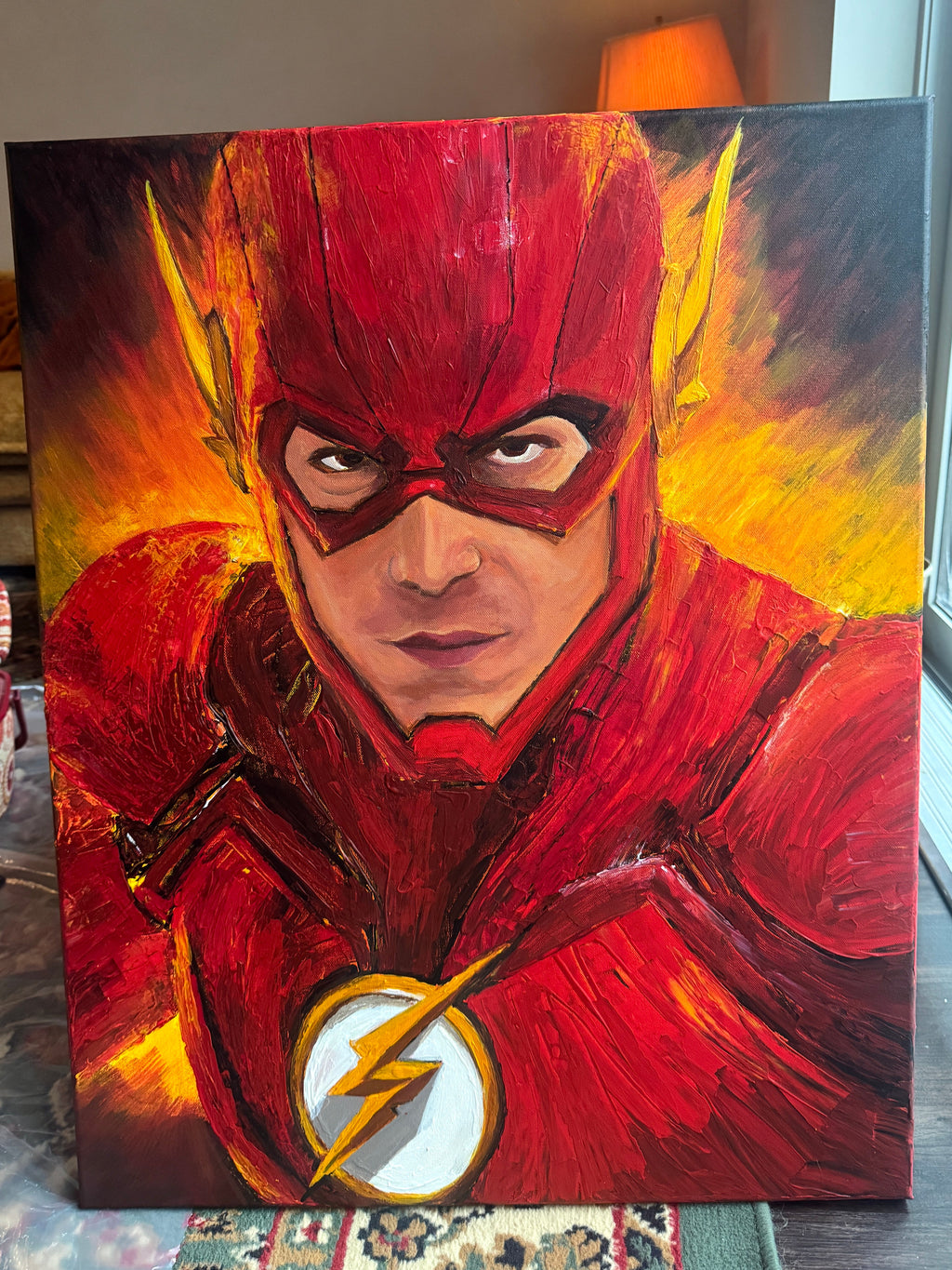 A square canvas painting of The Flash, featuring a textured, impasto-style portrait of the superhero in his iconic red suit with yellow lightning accents and a white lightning emblem on the chest, set against a fiery yellow and orange background. The canvas is shown standing on a patterned rug on a wooden floor in a home interior.