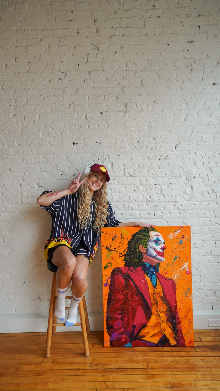 A woman sitting on a wooden stool next to a large canvas painting of the Joker on an orange background, posed in front of a white brick wall on a wooden floor.